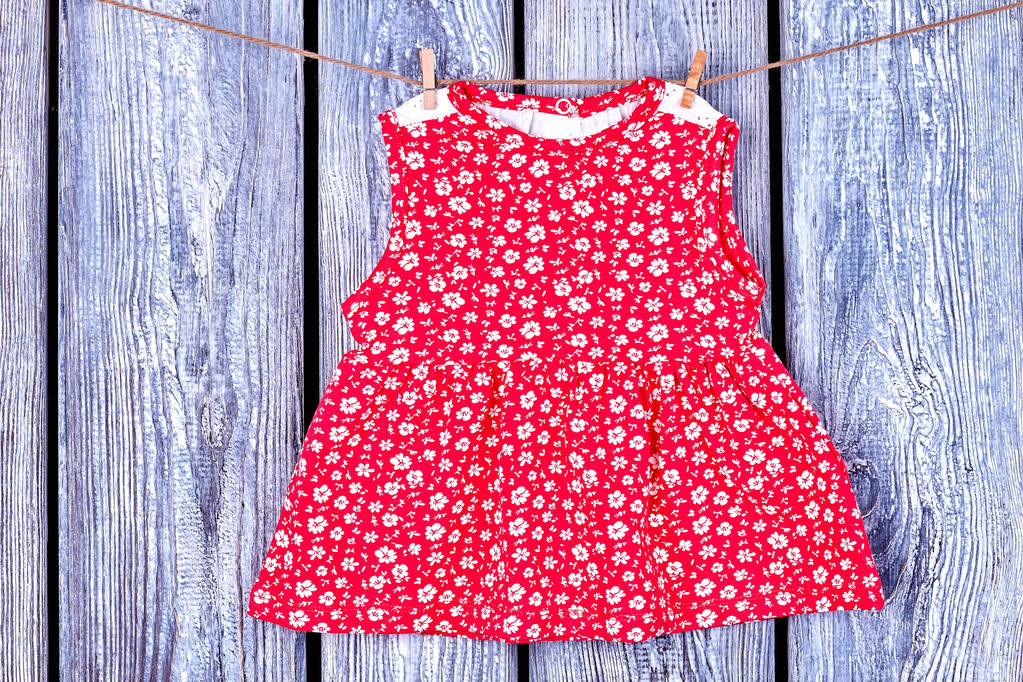 Baby red dress hanging on clothesline. Toddler girl patterned top drying on rope on vintage wooden background. Wet kids apparel in laundry.